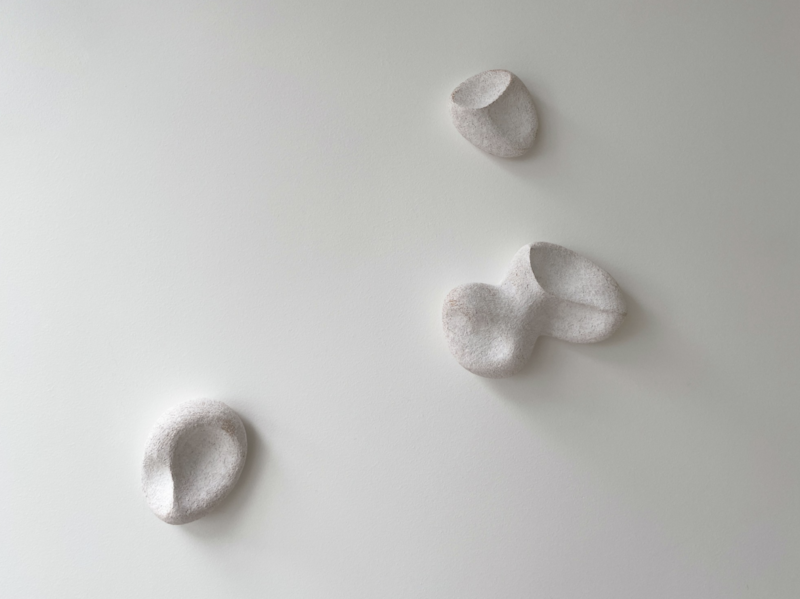 Yasha Butler’s “Pergamon No. 54, 2024” is a minimalist ceramic installation featuring organic, pebble-like forms arranged on a white wall. Each form varies in size and contour, creating a harmonious interplay of soft shadows and subtle textures.